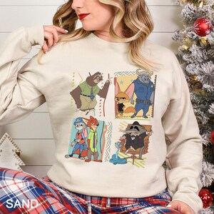 May include: Sand-colored sweatshirt featuring a colorful graphic of characters from the animated movie Zootopia. The design includes a sloth, fox, buffalo, rabbit, and raccoon. The sweatshirt has long sleeves and a crew neck.