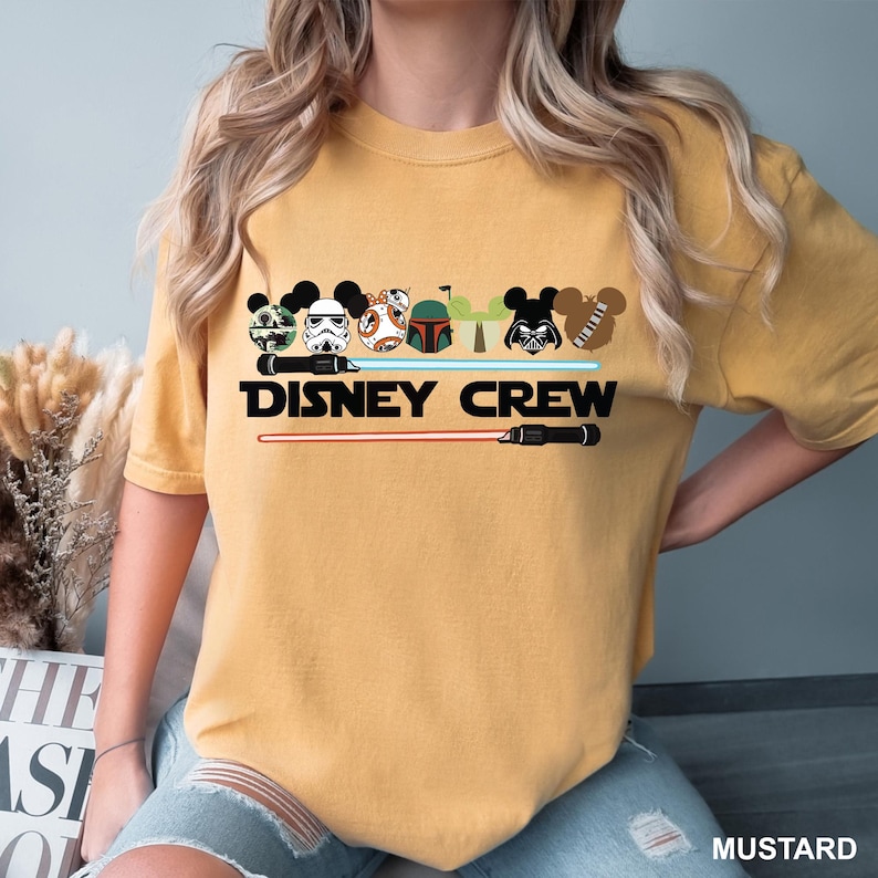 Pu&ograve; includere: Una maglietta gialla con una grafica di vari personaggi Disney e Star Wars che indossano le orecchie di Topolino. Il testo "DISNEY CREW" &egrave; stampato sotto la grafica.
