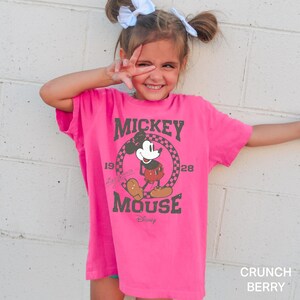May include: A pink t-shirt featuring a vintage Mickey Mouse graphic. The graphic depicts Mickey Mouse in black and white with a red shirt and black shoes. The text "Mickey Mouse" is in a black and white distressed font with the years "1928" and "1928" on either side of the graphic. The text "Disney" is in a smaller font below the graphic.