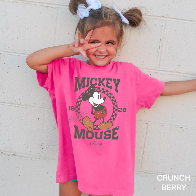 May include: A pink t-shirt featuring a vintage Mickey Mouse graphic. The graphic depicts Mickey Mouse in black and white with a red shirt and black shoes. The text "Mickey Mouse" is in a black and white distressed font with the years "1928" and "1928" on either side of the graphic. The text "Disney" is in a smaller font below the graphic.