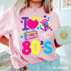 May include: Pink t-shirt with a colorful "I Love the 80's" graphic. The design includes a heart, sunglasses, Rubik's Cube, and a boombox. A white mug with a smiley face is held in the hand.