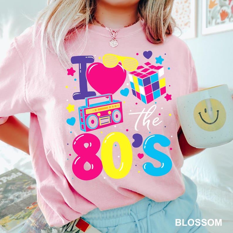 May include: Pink t-shirt with a colorful "I Love the 80's" graphic. The design includes a heart, sunglasses, Rubik's Cube, and a boombox. A white mug with a smiley face is held in the hand.