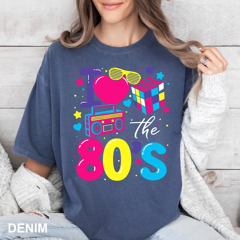 May include: Denim blue t-shirt with a colorful graphic that says "I love the 80s." The design includes a pink heart, yellow sunglasses, a Rubik's Cube, and a boombox. The word "DENIM" is at the bottom.