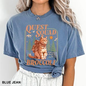 May include: Blue jean colored t-shirt with a graphic featuring a cat wearing a backpack, the words "QUEST SQUAD" and "BROCCOLI". The graphic also includes trees, a sun, and the text "No One Wants It, But It's Good For You."