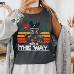 May include: Dark gray tank top featuring a graphic of a Mandalorian character with Mickey Mouse ears holding a heart-shaped balloon and Grogu. The design is set against a sunset background with the text "THIS IS THE WAY" in a white box.