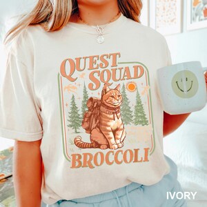 May include: Ivory-colored t-shirt featuring a graphic of a cat with a backpack, the words "QUEST SQUAD BROCCOLI", and a forest scene. A white mug with a smiley face is held in the hand. The shirt is a light cream color.