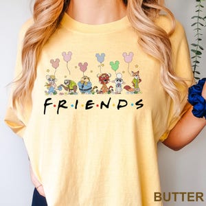 May include: Pale yellow t-shirt featuring the word "FRIENDS" in black, with coloured dots. Above the word are cartoon characters holding Mickey Mouse-shaped balloons. The shirt has the word "BUTTER" printed at the bottom right.