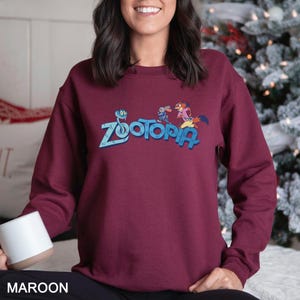 Puede incluir: Sudadera granate con la palabra "Zootopia" en azul y personajes de dibujos animados. La sudadera es de color granate liso. La palabra "MAROON" est&aacute; impresa en la parte inferior de la imagen.