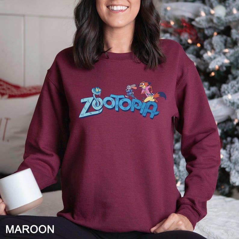 Puede incluir: Sudadera granate con la palabra "Zootopia" en azul y personajes de dibujos animados. La sudadera es de color granate liso. La palabra "MAROON" est&aacute; impresa en la parte inferior de la imagen.