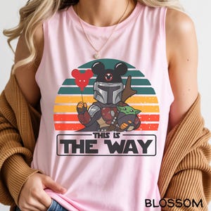May include: Pink tank top featuring a cartoon design with a Mandalorian character, Baby Yoda, and a Mickey Mouse head holding a heart-shaped balloon. The text "THIS IS THE WAY" is displayed below the characters, with a sunset background.