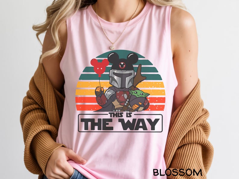 May include: Pink tank top featuring a cartoon design with a Mandalorian character, Baby Yoda, and a Mickey Mouse head holding a heart-shaped balloon. The text "THIS IS THE WAY" is displayed below the characters, with a sunset background.