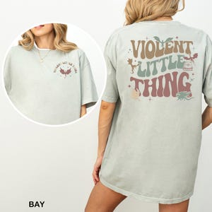 May include: A light green t-shirt with the text "VIOLENT LITTLE THING" in brown, green, and pink. The shirt has a graphic of a dragon and other small illustrations. The front of the shirt has the text "VIOLENT LITTLE THING" and a dragon illustration.