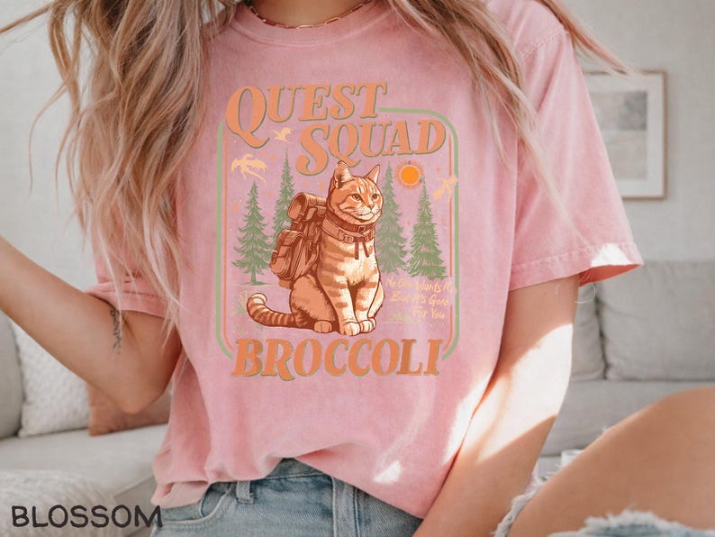 May include: Pink t-shirt with a graphic featuring a cat wearing a backpack, the words "QUEST SQUAD BROCCOLI", and a forest scene. The shirt has a crew neck and short sleeves.