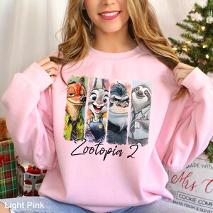 May include: Light pink sweatshirt featuring the characters from Zootopia 2. The design includes Nick Wilde, Judy Hopps, Chief Bogo, and Flash the sloth, with the text "Zootopia 2" in a script font.