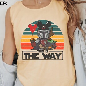 May include: Butter-colored tank top featuring a graphic of a Mandalorian-Mickey Mouse hybrid holding a red heart balloon and Grogu. The design is set against a sunset backdrop with the text "THIS IS THE WAY" in a black rectangle.