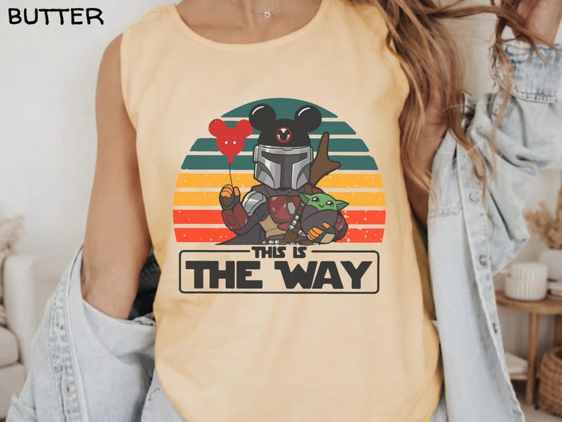 May include: Butter-colored tank top featuring a graphic of a Mandalorian-Mickey Mouse hybrid holding a red heart balloon and Grogu. The design is set against a sunset backdrop with the text "THIS IS THE WAY" in a black rectangle.