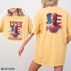 May include: A mustard yellow short-sleeve t-shirt featuring a graphic of Spider-Man in front of an American flag. The back shows a large Spider-Man print, while the front inset displays the text 1776 UNITED STATES INDEPENDENCE DAY.