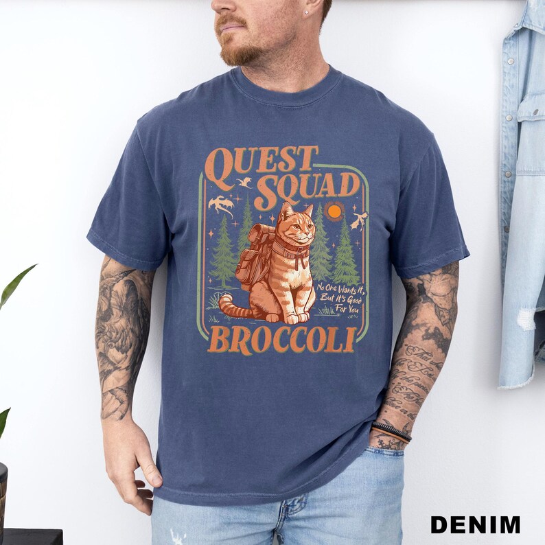 May include: A navy blue t-shirt with a graphic featuring a cat wearing a backpack, the words "QUEST SQUAD BROCCOLI", and the phrase "No One Wants It, But It's Good For You".