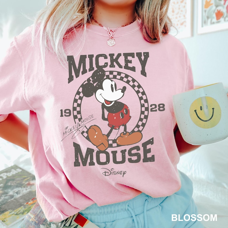 May include: Pink t-shirt featuring a vintage Mickey Mouse graphic. The graphic shows Mickey Mouse in a red and black outfit, standing in a checkered circle with the text "Mickey Mouse" and "Disney" below. The text "1928" is on the right side of the circle and "Mickey Mouse" is written in cursive on the left side of the circle.