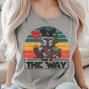 May include: Gray t-shirt featuring a graphic of a Mandalorian character with a Mickey Mouse head, holding a red balloon and Baby Yoda. The design is set against a retro sunset with the text "THIS IS THE WAY" in a rectangular box.