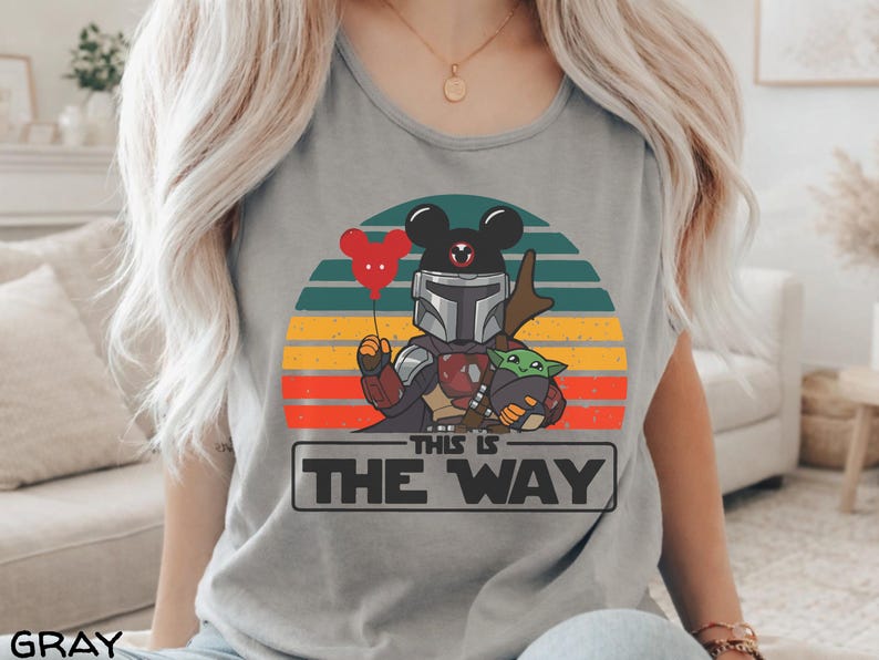 May include: Gray t-shirt featuring a graphic of a Mandalorian character with a Mickey Mouse head, holding a red balloon and Baby Yoda. The design is set against a retro sunset with the text "THIS IS THE WAY" in a rectangular box.