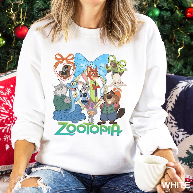 Op de afbeelding: Witte sweatshirt met een kleurrijke afbeelding van personages uit de animatiefilm "Zootopia". Het ontwerp bevat een blauwe strik, een gouden lijst en het woord "Zootopia" in groen en blauw. De sweatshirt heeft een ronde hals.