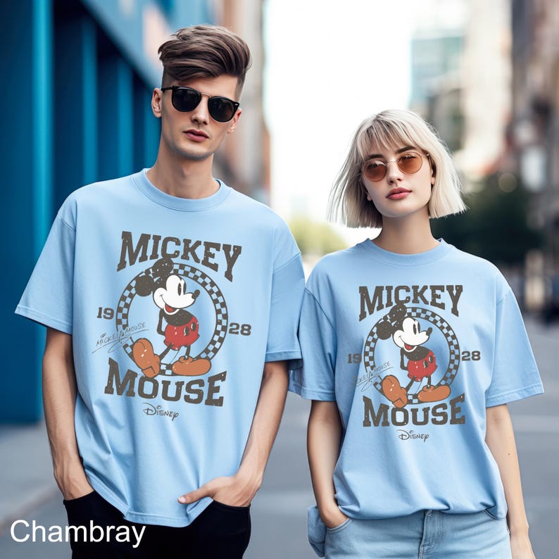 May include: Two light blue t-shirts featuring a vintage Mickey Mouse graphic. The graphic depicts Mickey Mouse in a red and black outfit, standing within a checkered circle with the text "Mickey Mouse" and "1928" above and below the circle. The text "Disney" is positioned beneath the circle.