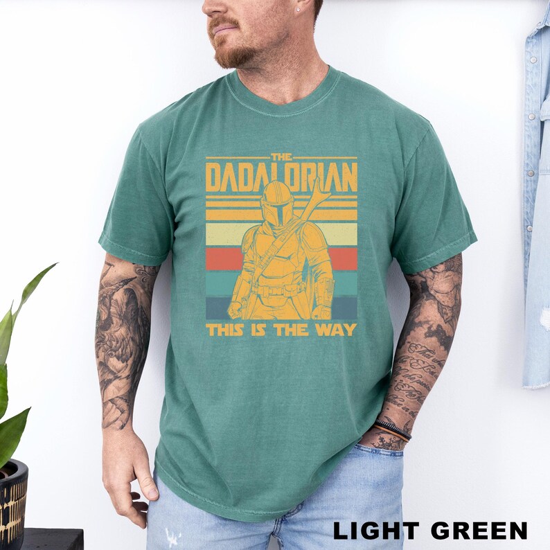 May include: Light green t-shirt featuring a graphic of a character with a helmet and weapon, with the text "THE DADALORIAN" above and "THIS IS THE WAY" below. The design has retro stripes in orange, yellow, and teal.