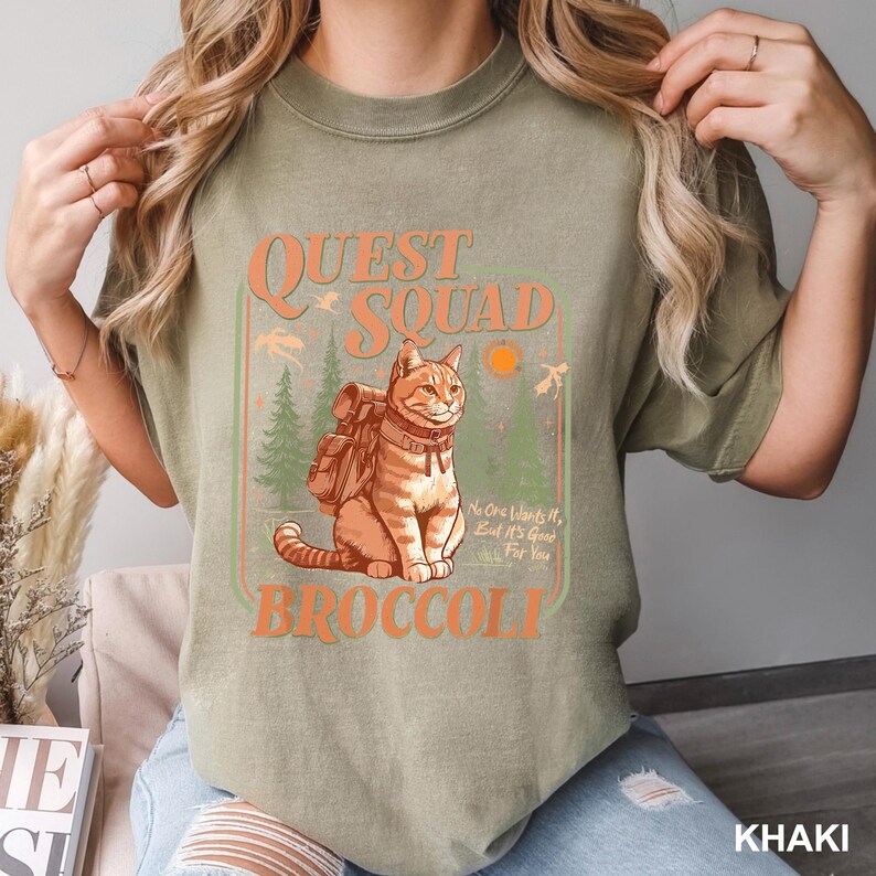 May include: Khaki-colored t-shirt with a graphic featuring a cat wearing a backpack, the words "QUEST SQUAD BROCCOLI", and the phrase "No One Wants It, But It's Good For You". The design includes trees and a sun.