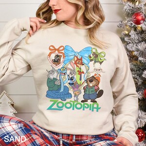 Op de afbeelding: Een zandkleurige sweatshirt met een kleurrijke afbeelding van personages uit de animatiefilm "Zootopia". Het ontwerp bevat een blauwe strik, een hartvormige lijst en het woord "Zootopia" in het groen. De sweatshirt heeft lange mouwen.