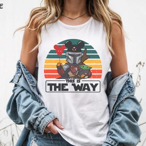 May include: White tank top featuring a graphic of a Mandalorian-inspired character with a Mickey Mouse head, holding a heart-shaped balloon, and Grogu. The design is set against a sunset backdrop with the text "THIS IS THE WAY".