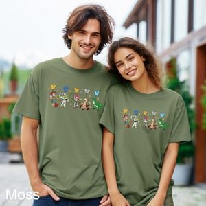 K&ouml;nnte beinhalten: Zwei Personen tragen gr&uuml;ne T-Shirts mit einem Aufdruck von Toy Story-Charakteren, die Ballons halten. Zu den Charakteren geh&ouml;ren Woody, Buzz Lightyear, Jessie, Bullseye und Rex.