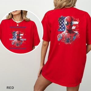 May include: A bright red t-shirt featuring a graphic of the superhero Spider-Man centered over a stylized American flag. The design includes the text UNITED STATES. The back of the shirt displays a larger version of the same graphic print.