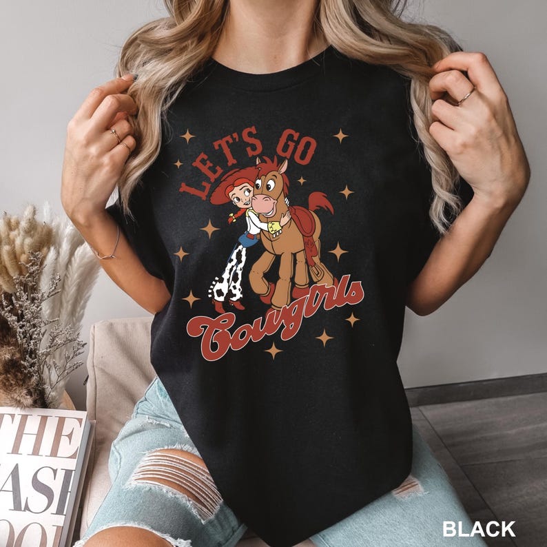 May include: Black t-shirt featuring a cartoon cowgirl riding a horse. The text "Let's Go Cowgirls" is printed on the shirt.