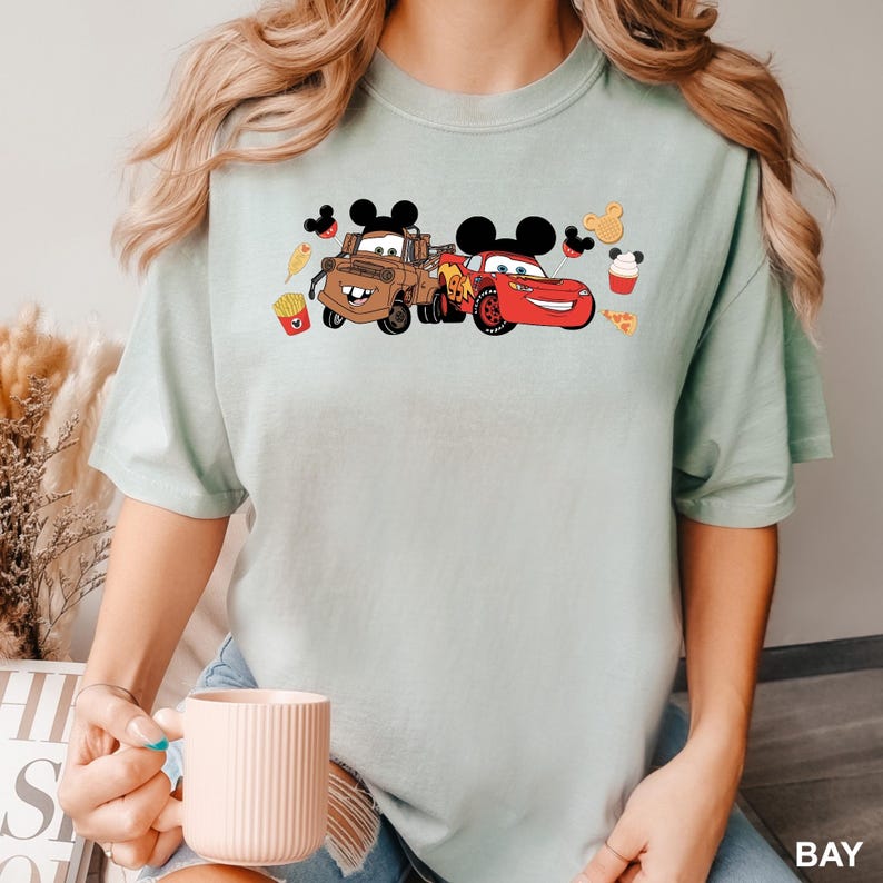 May include: A light blue t-shirt with a graphic of Mickey Mouse ears, Mater and Lightning McQueen from Cars. The graphic also includes Mickey Mouse ears, a hot dog, fries, pizza, and a cupcake.