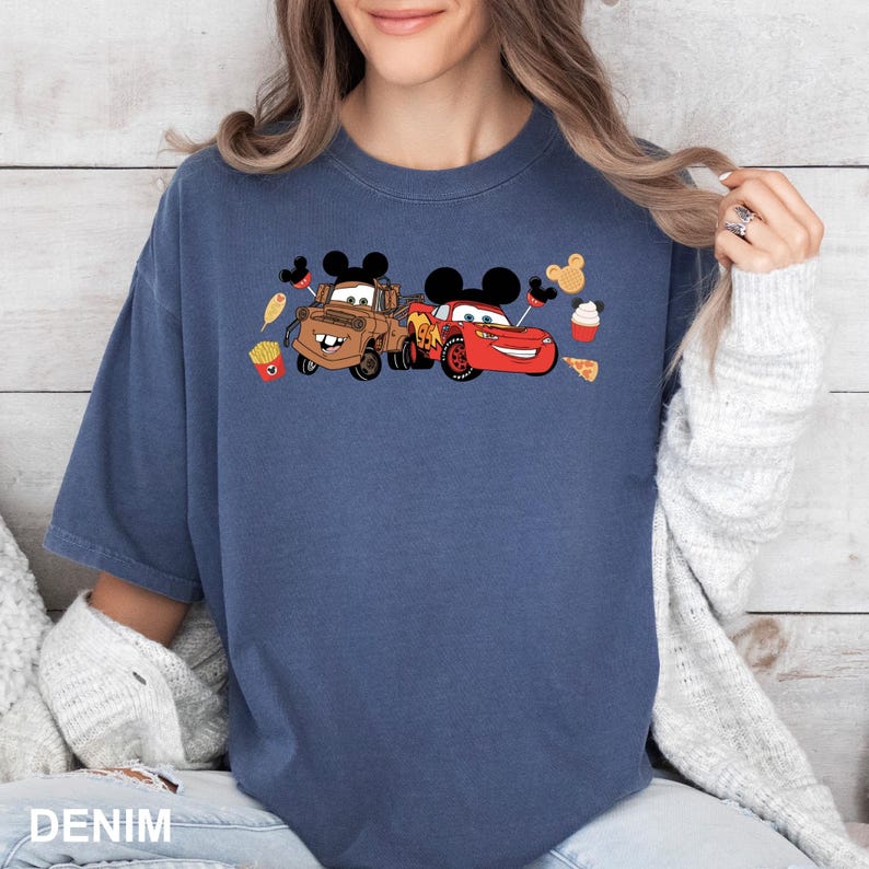 May include: A blue t-shirt with a graphic of Mater and Lightning McQueen from Cars, wearing Mickey Mouse ears and surrounded by Mickey Mouse ears and food items.