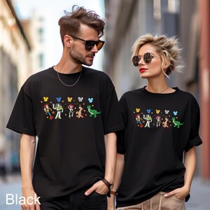 K&ouml;nnte beinhalten: Zwei Personen tragen schwarze T-Shirts mit einem bunten Grafikdruck von Toy Story-Charakteren und Mickey-Maus-Ohren. Die Grafik enth&auml;lt Woody, Buzz Lightyear, Jessie, Rex und einen gr&uuml;nen Au&szlig;erirdischen.