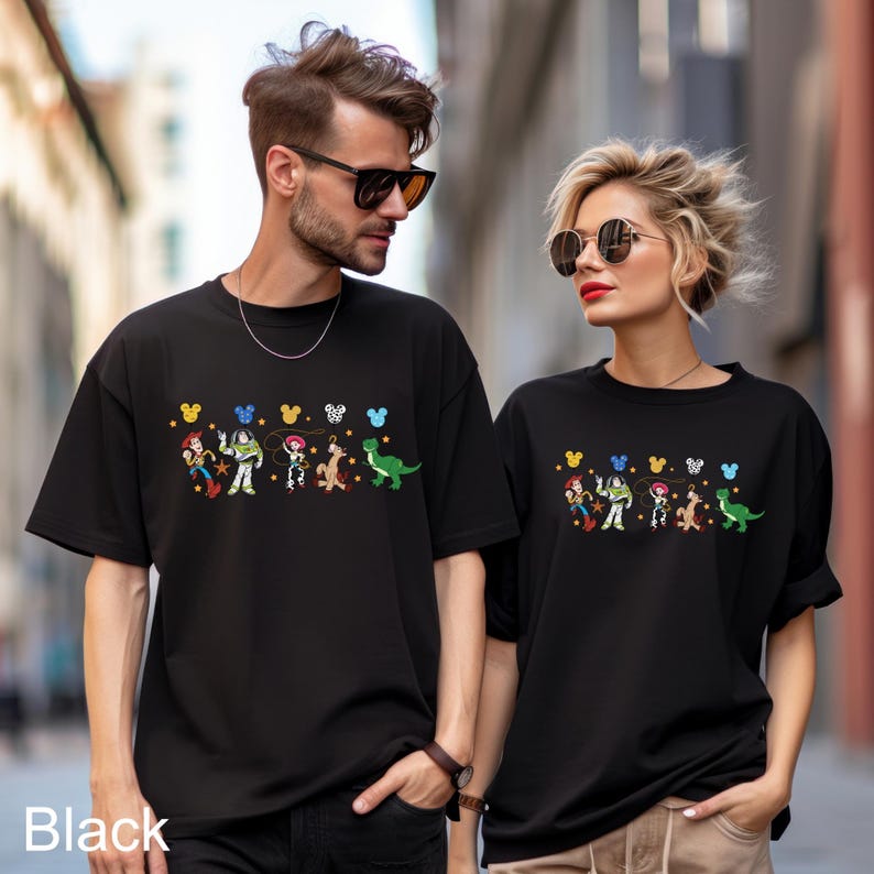 K&ouml;nnte beinhalten: Zwei Personen tragen schwarze T-Shirts mit einem bunten Grafikdruck von Toy Story-Charakteren und Mickey-Maus-Ohren. Die Grafik enth&auml;lt Woody, Buzz Lightyear, Jessie, Rex und einen gr&uuml;nen Au&szlig;erirdischen.