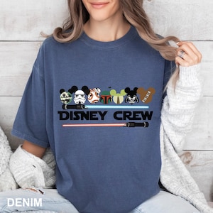 Pu&ograve; includere: Una t-shirt blu denim con una grafica di personaggi di Star Wars che indossano le orecchie di Topolino. Il testo "DISNEY CREW" &egrave; stampato sotto la grafica. La grafica include anche due spade laser rosse.