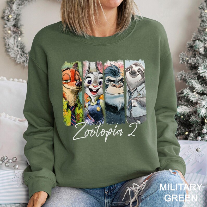 May include: A military green sweatshirt featuring a colorful graphic of characters from the animated movie Zootopia. The graphic includes Nick Wilde, Judy Hopps, Chief Bogo, and Flash. The text "Zootopia 2" is written in white script below the characters.