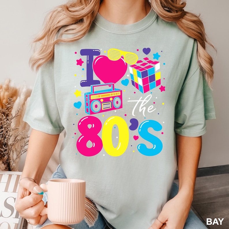 May include: A light green t-shirt with a colorful graphic that says "I love the 80's". The design includes a pink heart, a Rubik's cube, a boombox, and other 80s-themed elements.