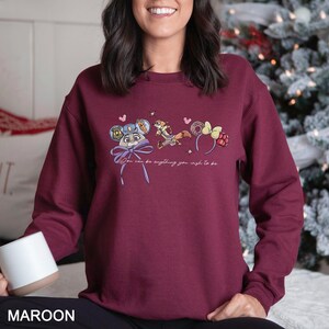 May include: Maroon sweatshirt featuring a whimsical illustration of cartoon character ears and a bow. The text "You can be anything you wish to be" is printed below the graphic. The sweatshirt is a solid maroon color.