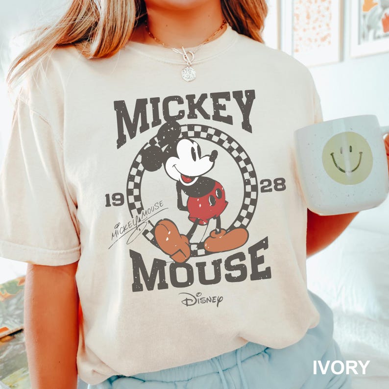 May include: A white t-shirt featuring a vintage-style graphic of Mickey Mouse. The graphic depicts Mickey Mouse in a red and black outfit, standing within a circle that includes the words "Mickey Mouse" and the numbers "19" and "28". The text "Disney" is also part of the graphic.