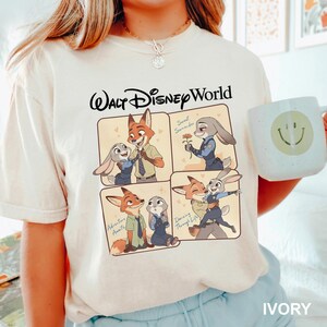 以下が含まれることがあります： 映画「ズートピア」のキャラクターが描かれたアイボリー色のTシャツ。「ウォルト・ディズニー・ワールド」の文字と、様々なポーズのキャラクターが描かれたパネルがデザインされています。白いマグカップにはスマイルマークが描かれています。