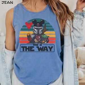 May include: Blue tank top featuring a graphic of a Mandalorian character with a Mickey Mouse head, holding a red heart-shaped balloon, and Grogu. The design is set against a sunset backdrop with the text "THIS IS THE WAY".