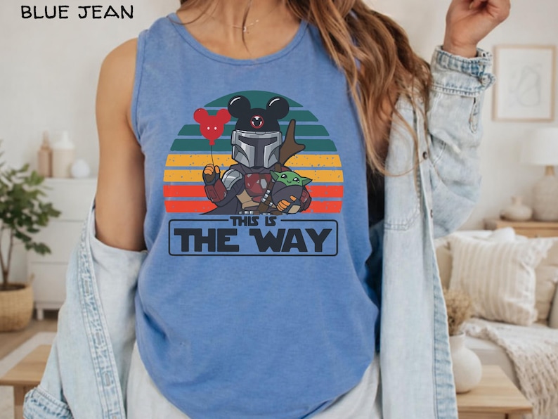 May include: Blue tank top featuring a graphic of a Mandalorian character with a Mickey Mouse head, holding a red heart-shaped balloon, and Grogu. The design is set against a sunset backdrop with the text "THIS IS THE WAY".
