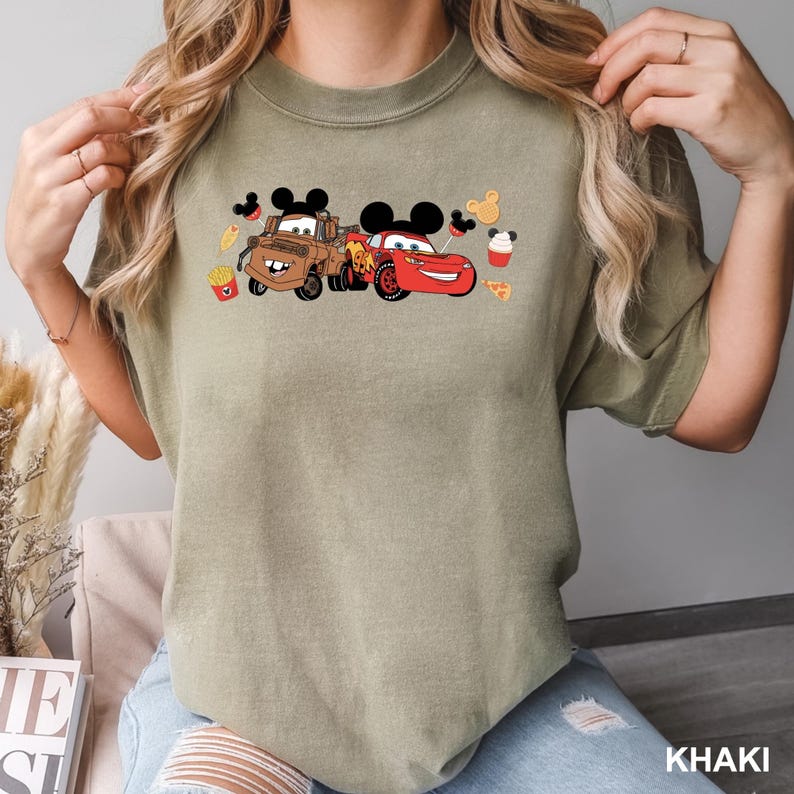 May include: A khaki t-shirt with a graphic print of two cartoon cars wearing Mickey Mouse ears. The cars are surrounded by cartoon food items, including Mickey Mouse shaped pretzels, french fries, and pizza slices.