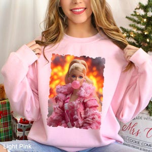 May include: Light pink sweatshirt featuring a Barbie doll graphic blowing a bubblegum bubble. The doll is wearing a pink fur coat and is set against a fiery background. The sweatshirt is a solid light pink color.