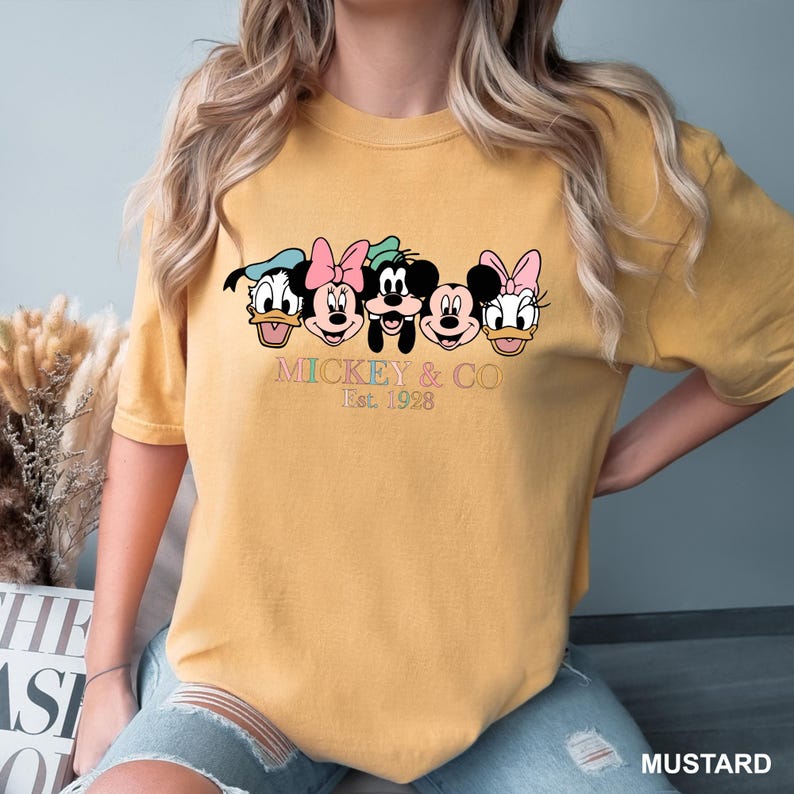 May include: A mustard yellow t-shirt with a graphic of Mickey Mouse and friends. The text "Mickey & Co. Est. 1928" is printed below the graphic.