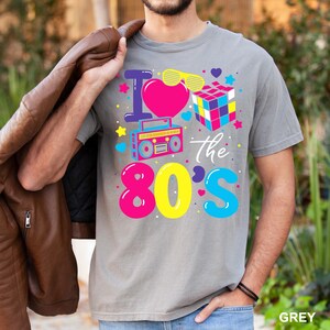May include: A grey t-shirt with a colorful graphic that says "I love the 80's". The design includes a pink heart, a Rubik's cube, a boombox, and sunglasses. The shirt is made of a soft material.